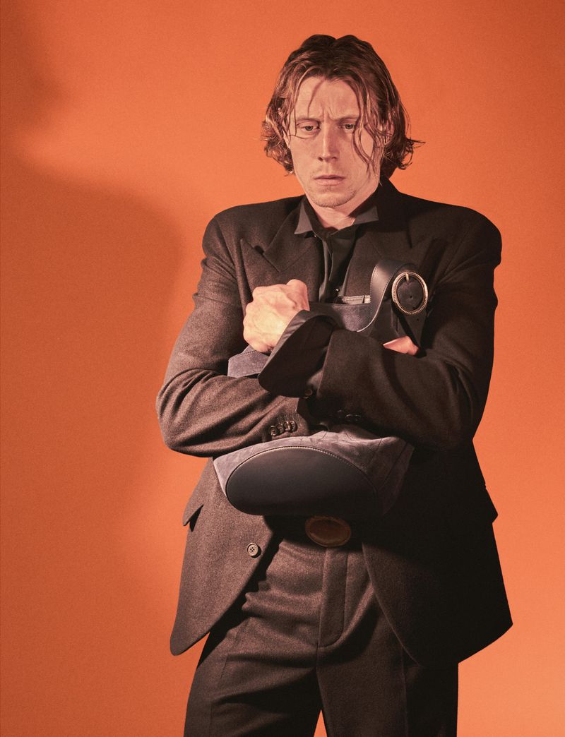 George MacKay wearing a dark suit against a solid warm orange background for HERO Magazine