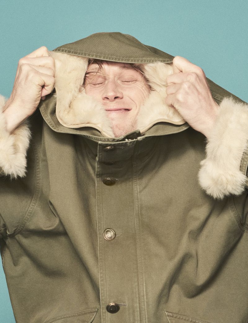George MacKay wearing an olive green hooded parka with a cream shearling lining against a teal blue background