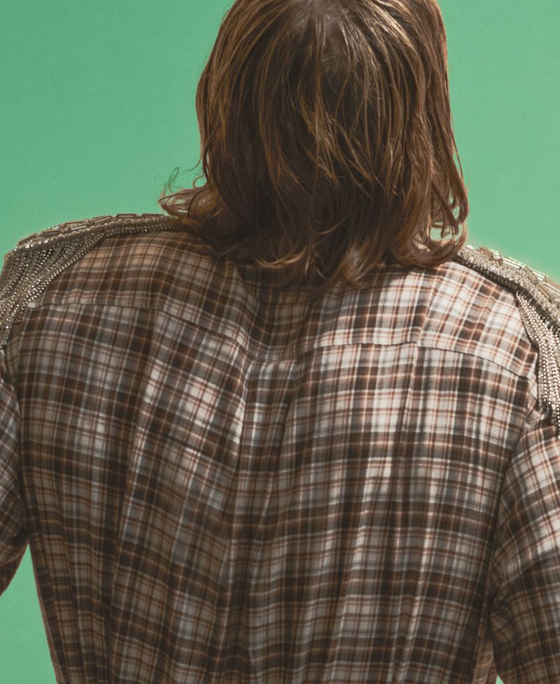 Rear view of George MacKay wearing a plaid shirt with embellished epaulets against a green backdrop.