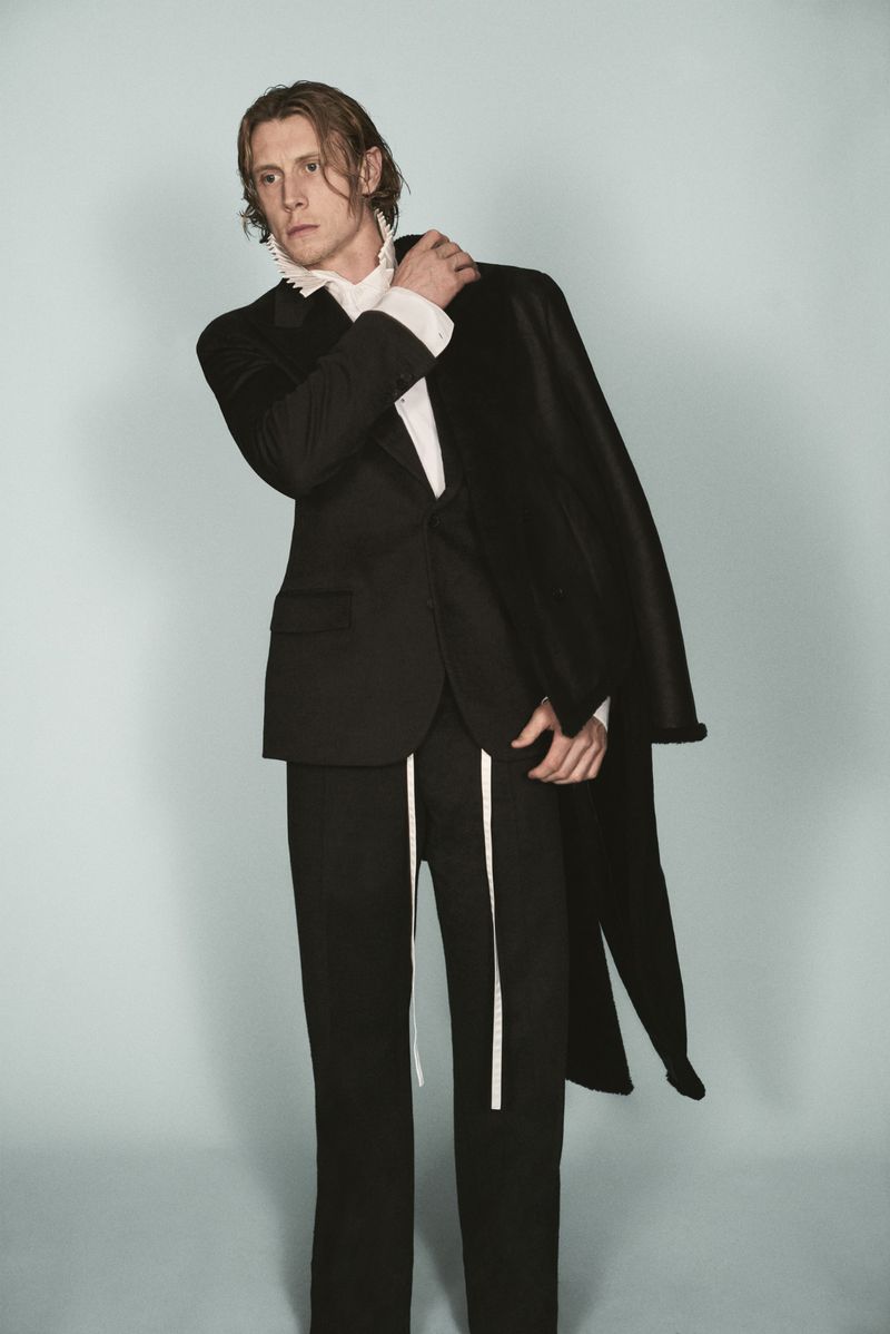 Actor George MacKay wearing a black suit and white ruffled shirt in front of a pale grey studio background for HERO Magazine.