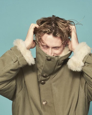 George MacKay pulling his hair while wearing an olive green coat with fur cuffs against a turquoise background