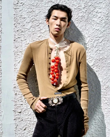 East Asian male model wearing a tan henley and black trousers standing against a textured white stucco wall with strong sunlight