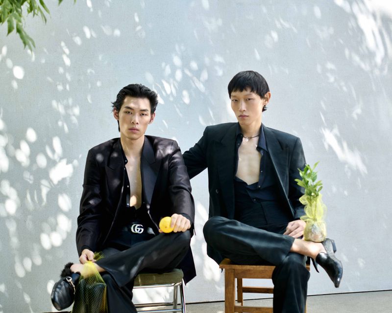 Two male models in dark suits seated on chairs outdoors against a plain wall with dappled leaf shadows.