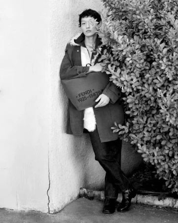 Monochrome fashion editorial portrait of a model leaning against a wall next to a leafy bush holding a Fendi bag