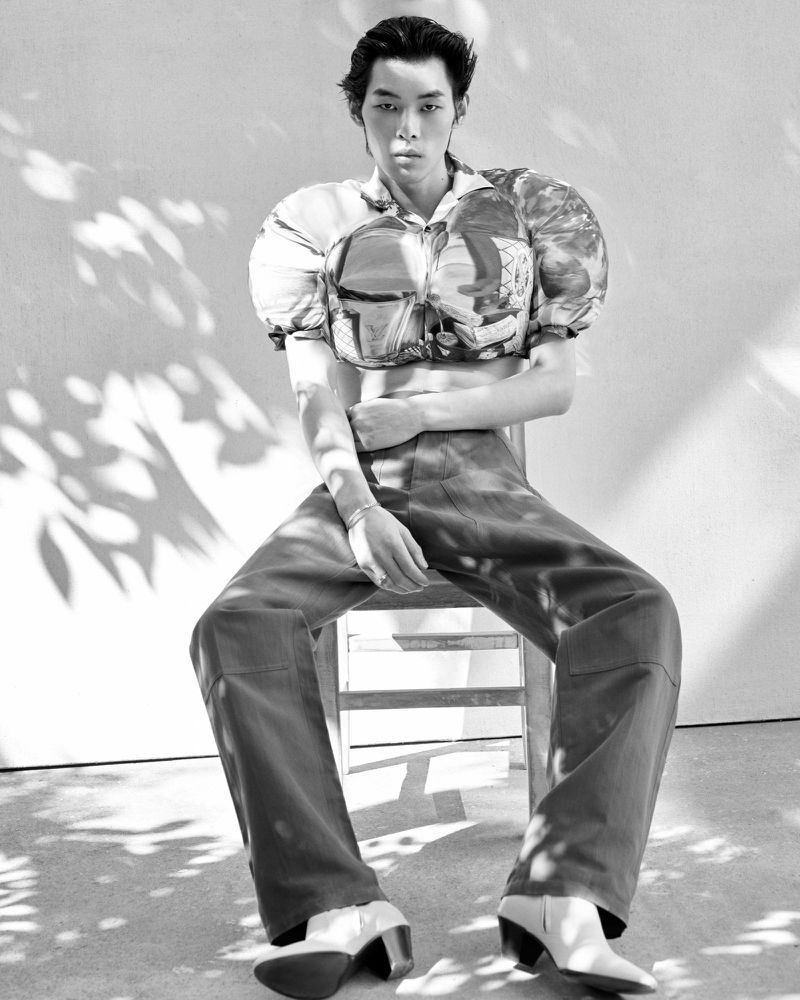 Monochrome fashion editorial portrait of a male model seated on a chair wearing a patterned crop top and trousers.