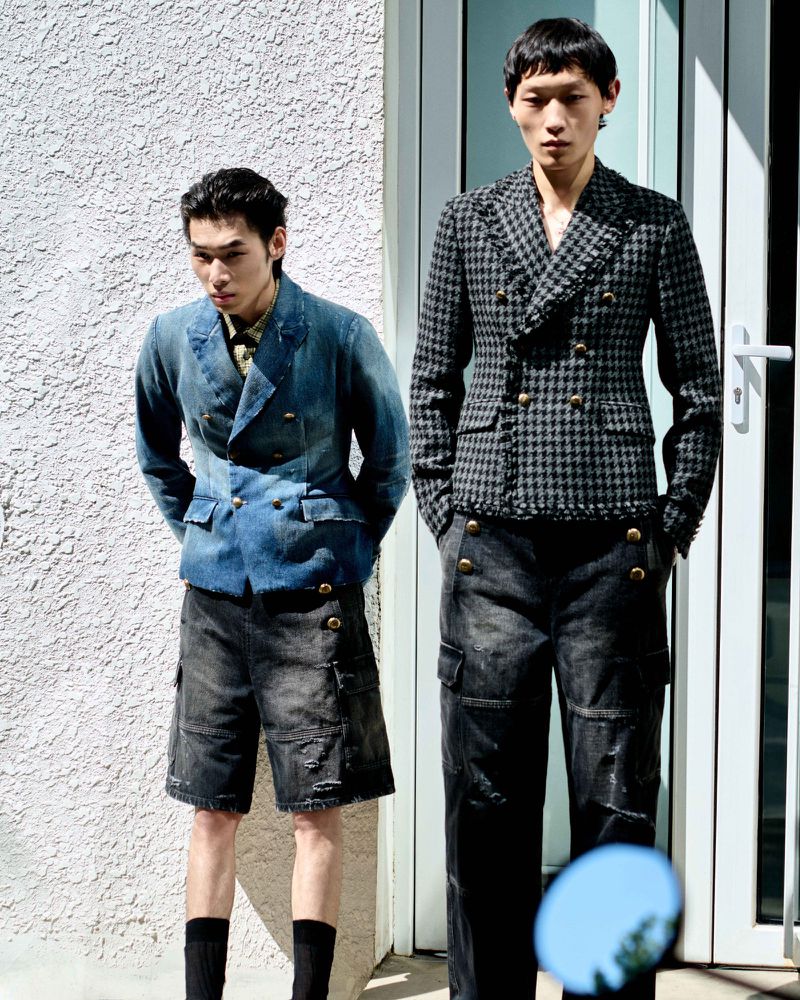 Two male models standing outdoors against a stucco wall and doorway wearing tailored jackets and dark trousers.