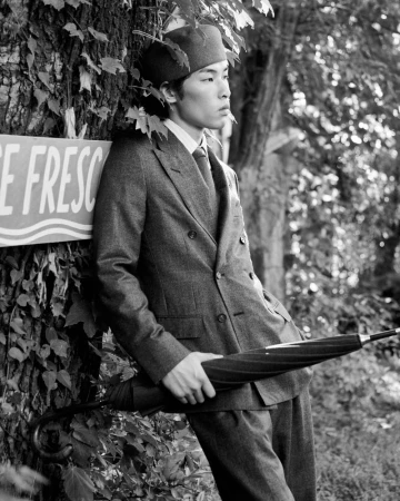 Man wearing a tailored grey suit and flat cap leaning against a tree trunk while holding a closed umbrella.