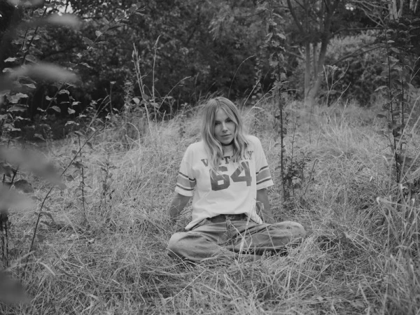 Sienna Miller seated cross-legged in a grassy field wearing a printed t-shirt and loose trousers in a monochrome portrait.