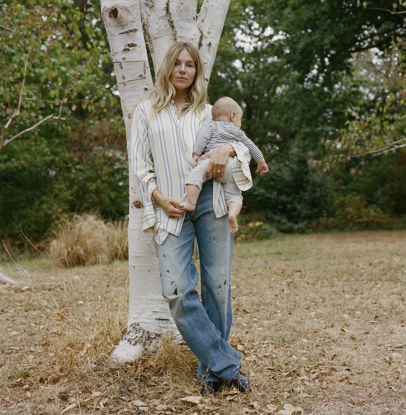 Sienna Miller holding an infant child while leaning against a white birch tree in an outdoor park setting.