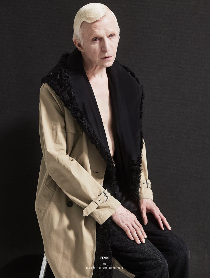 Man with platinum blonde hair wearing a beige coat with black fur collar seated against a dark background