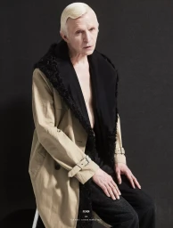 Man with platinum blonde hair wearing a beige coat with black fur collar seated against a dark background