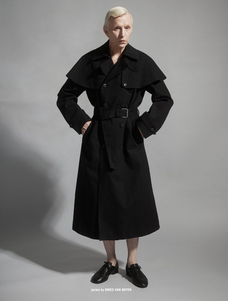 Man wearing a long black belted trench coat with cape shoulders and black shoes against a grey seamless background