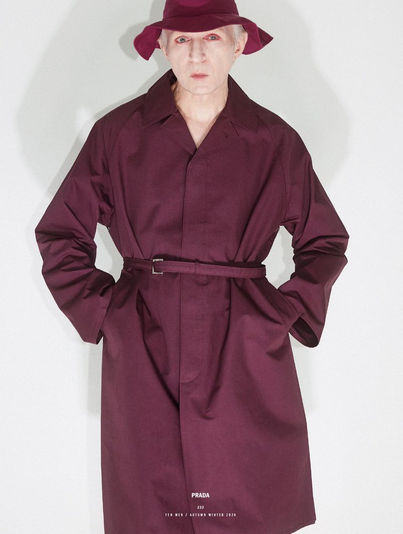 Man wearing burgundy trench coat and matching bucket hat against a plain light wall for 10 Men Magazine editorial