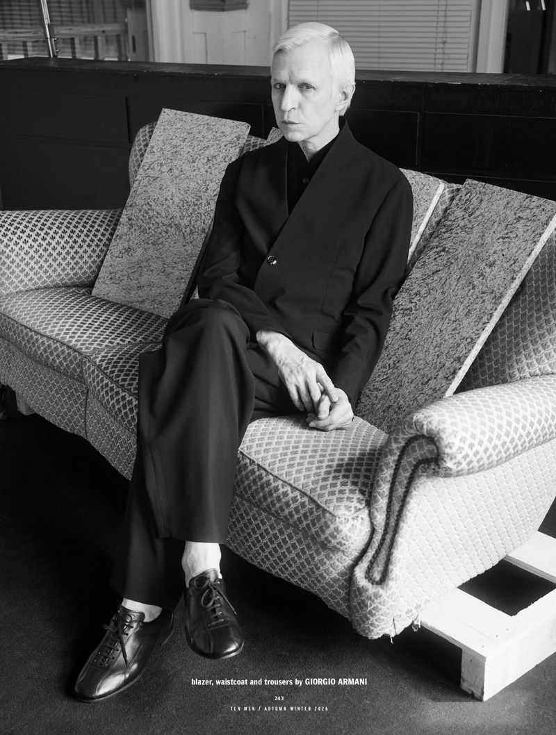 Monochrome portrait of a man seated on a patterned sofa wearing a dark suit jacket, trousers, and leather shoes