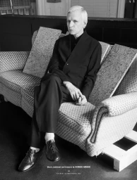 Monochrome portrait of a man seated on a patterned sofa wearing a dark suit jacket, trousers, and leather shoes