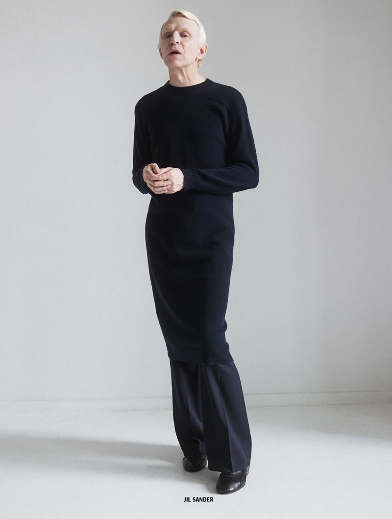 Man wearing a long black sweater and dark trousers standing against a white seamless background for 10 Men Magazine.