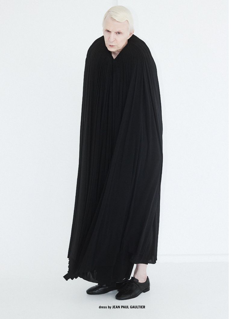 Full-length portrait of a man wearing a long pleated black cloak and black shoes against a white background for 10 Men Magazine.