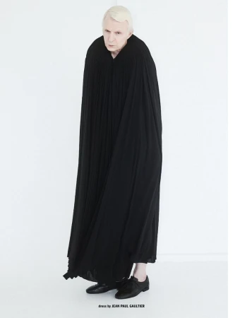 Full-length portrait of a man wearing a long pleated black cloak and black shoes against a white background for 10 Men Magazine.