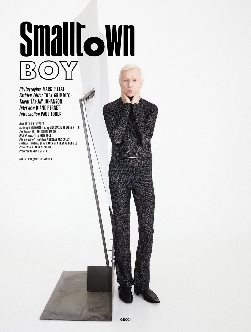 Blonde man wearing a black patterned turtleneck and trousers standing next to a metallic sculpture against a white background