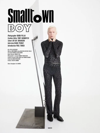 Blonde man wearing a black patterned turtleneck and trousers standing next to a metallic sculpture against a white background