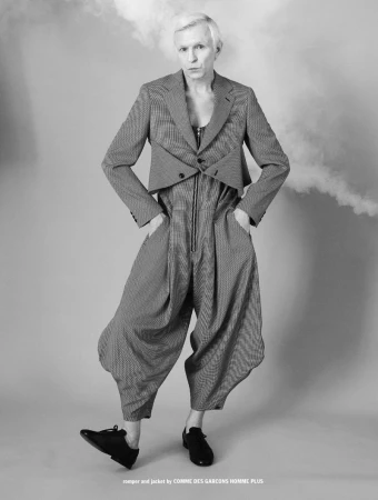Monochrome portrait of a man wearing a patterned tailored jacket and drop-crotch jumpsuit against a plain grey backdrop.