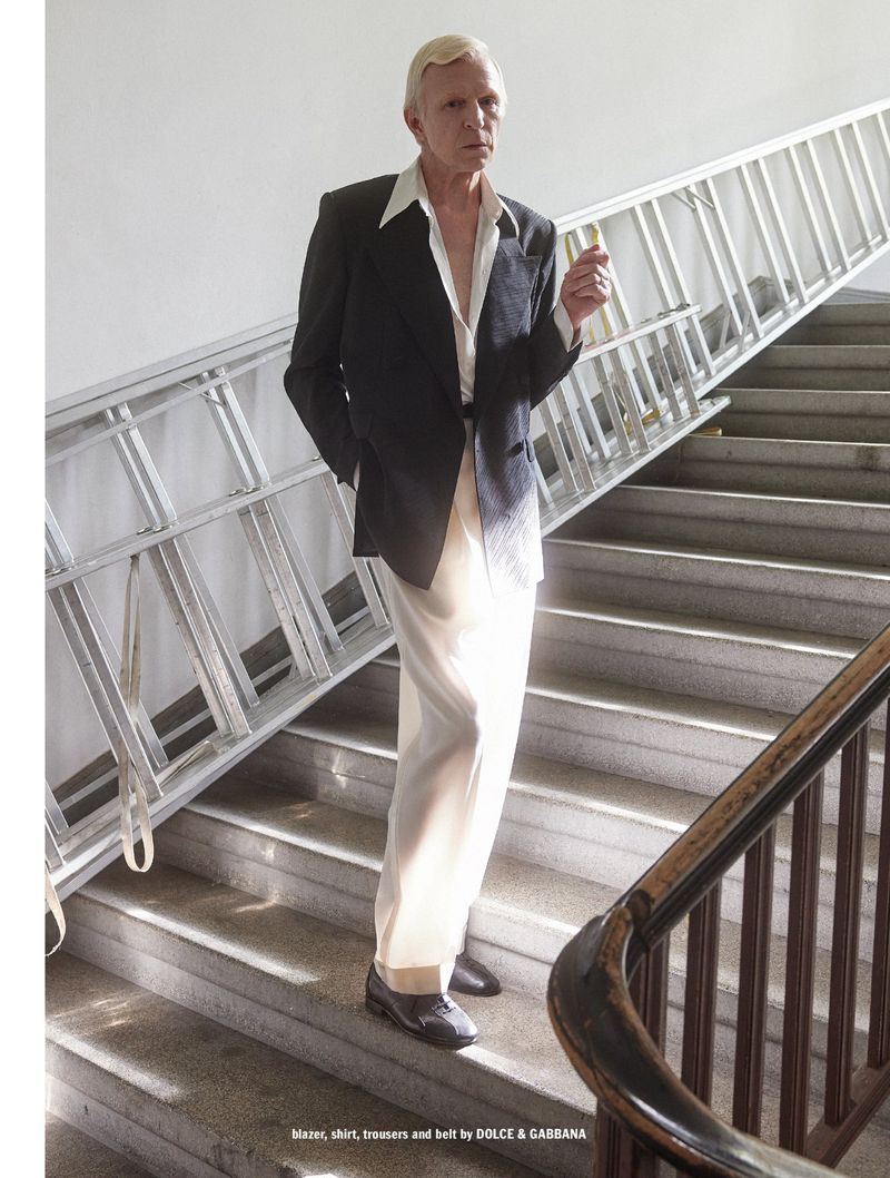 A man in a dark blazer, open white shirt, and light trousers standing on a grand indoor staircase next to a metal ladder.