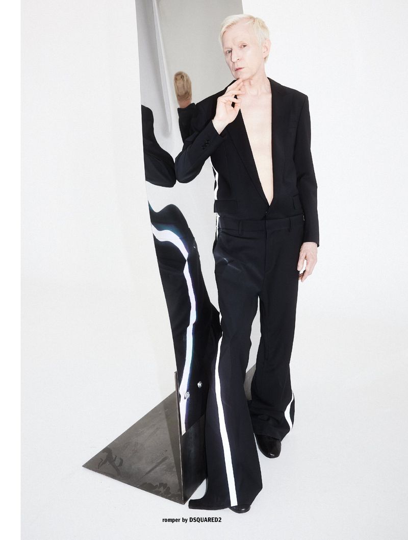 Blonde man wearing a black suit jacket and matching trousers posing next to a vertical mirror against a white studio wall