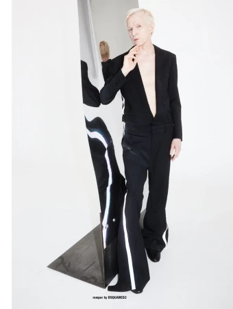 Blonde man wearing a black suit jacket and matching trousers posing next to a vertical mirror against a white studio wall