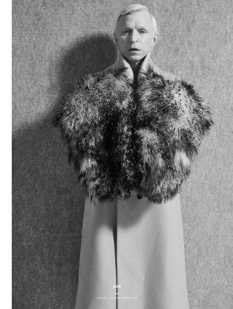 Man wearing a textured fur stole and long coat against a textured grey wall in a black and white editorial portrait.
