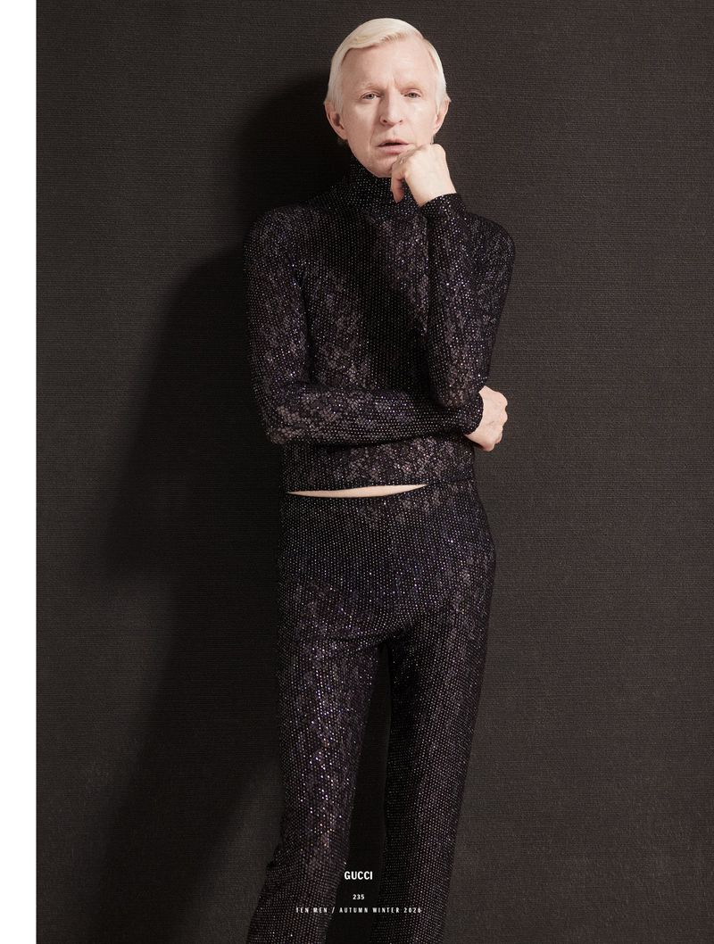 Man with blonde hair wearing a dark textured turtleneck and matching trousers against a dark background for 10 Men Magazine