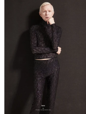 Man with blonde hair wearing a dark textured turtleneck and matching trousers against a dark background for 10 Men Magazine