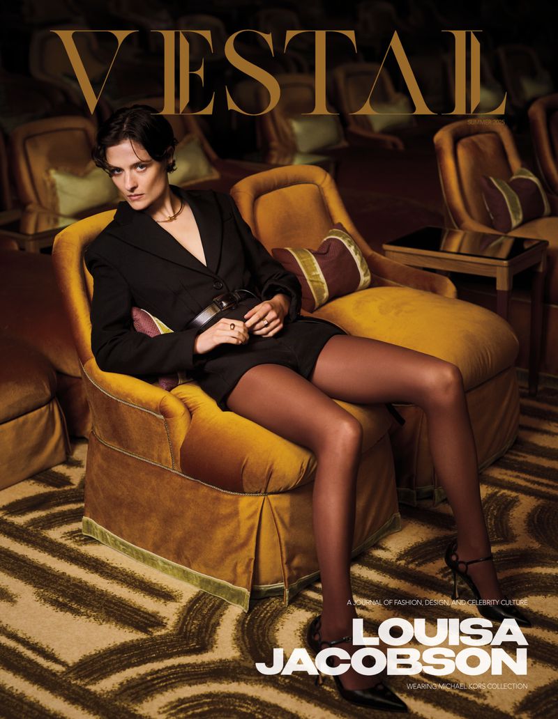 Magazine cover featuring a model in a black blazer and shorts sitting in a vintage chair in a theater-like setting with patterned carpet.