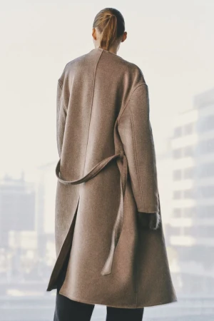 Model seen from behind wearing a long brown belted coat against a soft blurred background