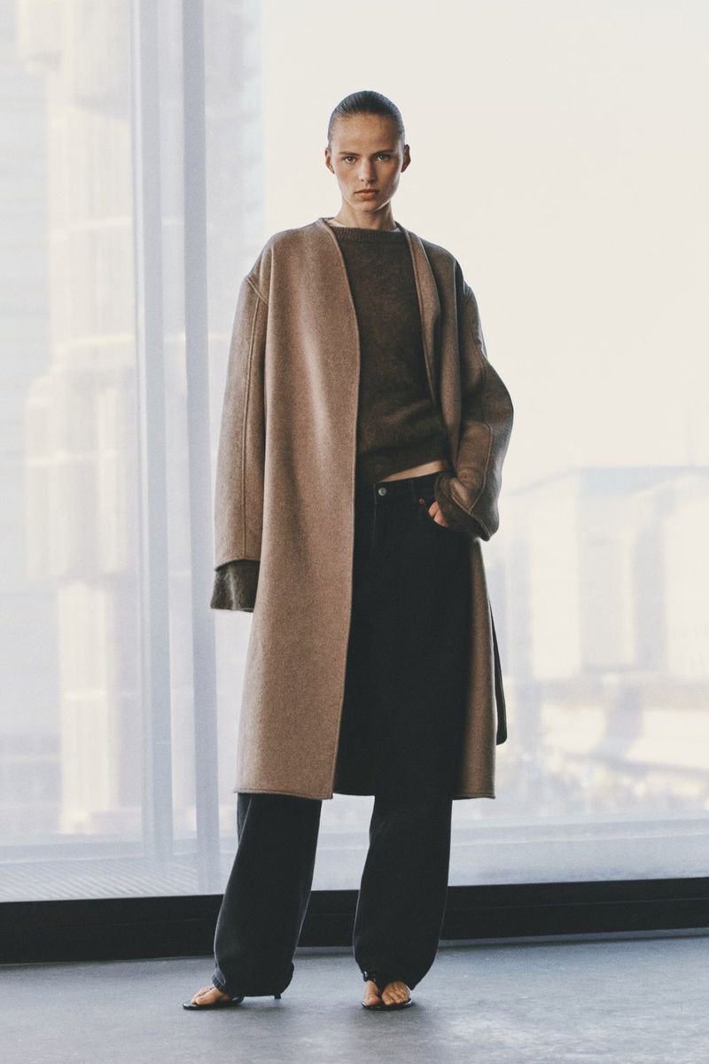 Model standing in a long camel coat and dark trousers against a large bright window