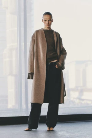 Model standing in a long camel coat and dark trousers against a large bright window