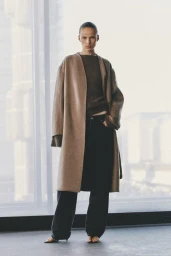 Model standing in a long camel coat and dark trousers against a large bright window