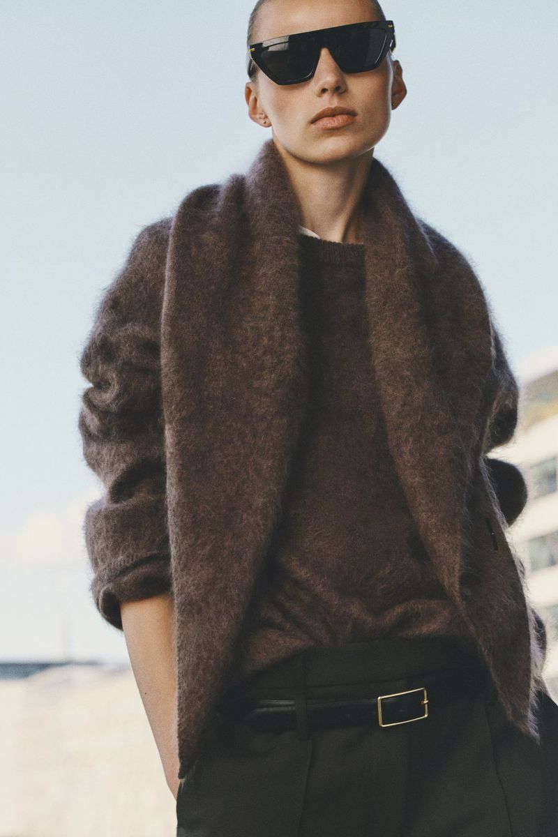 Model wearing dark sunglasses, brown fuzzy scarf, sweater, and dark trousers against an outdoor sky background.
