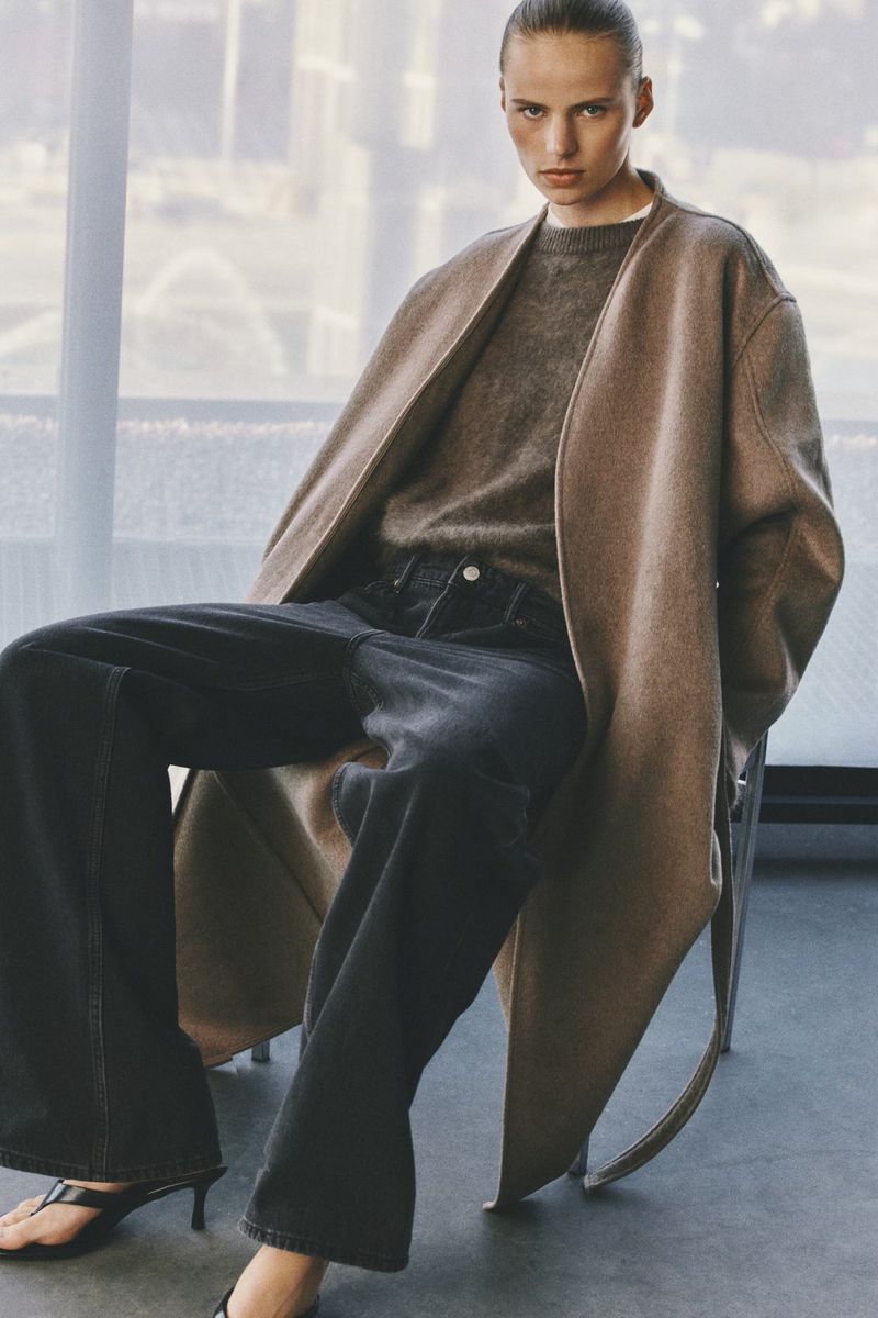 Model seated on a chair wearing a brown coat, grey knit sweater, and dark wide-leg trousers indoors.