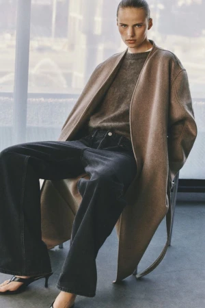 Model seated on a chair wearing a brown coat, grey knit sweater, and dark wide-leg trousers indoors.