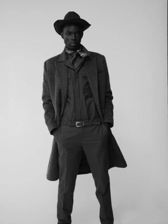 Model wearing a dark hat, patterned neck scarf, long coat, and button-up shirt standing against a neutral background.