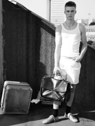 Monochrome portrait of a male model wearing a white tank top and shorts holding a large leather bag on a rooftop.