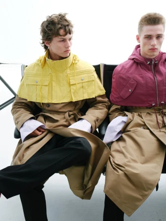 Two male models in tan trench coats seated against a white background for U Repubblica by Joseph Echenique.