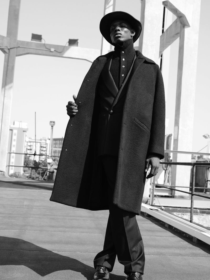 Black man wearing a dark overcoat, buttoned collar shirt, and wide-brimmed hat on a concrete rooftop.