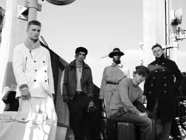 Group of five male models in layered outerwear posing outdoors near architectural structures in a black and white fashion editorial.