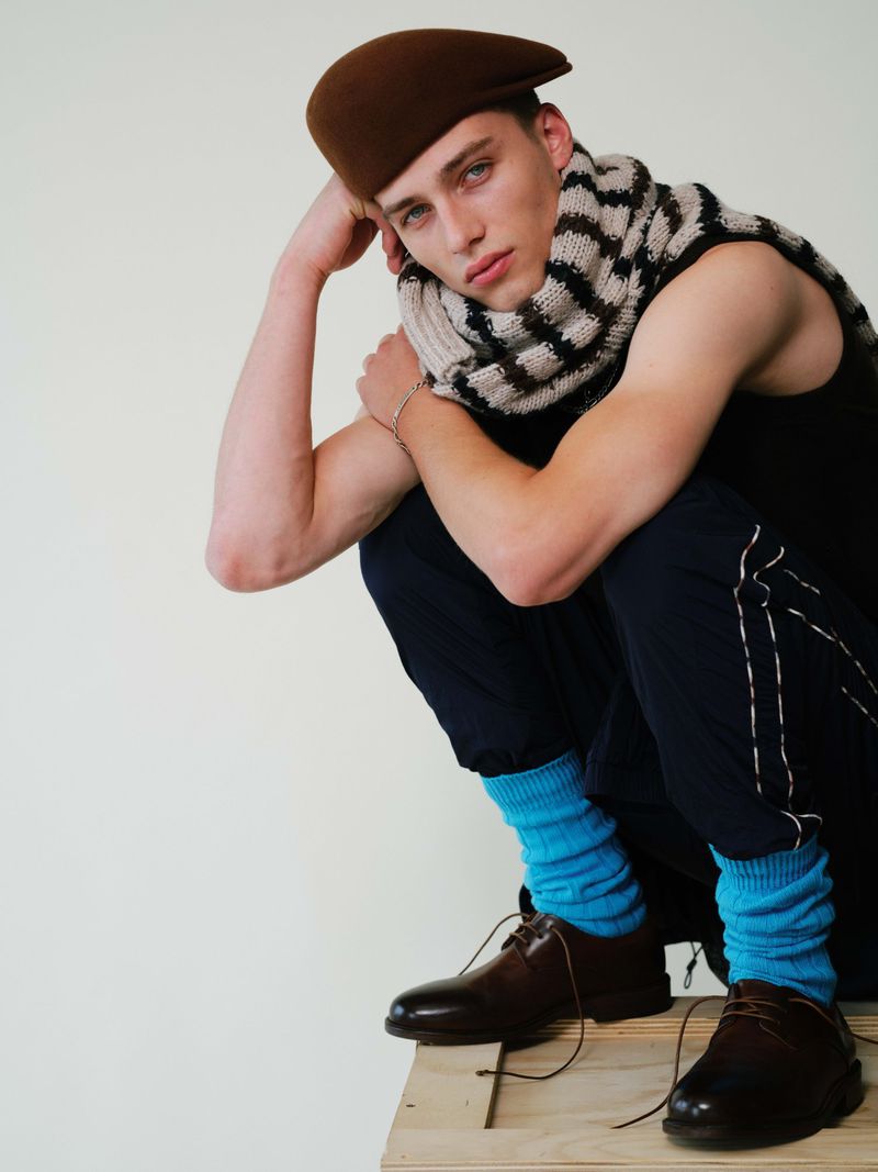 Model crouching wearing a brown beret, striped scarf, dark trousers, and bright blue socks against a plain light background