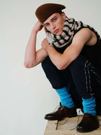 Model crouching wearing a brown beret, striped scarf, dark trousers, and bright blue socks against a plain light background