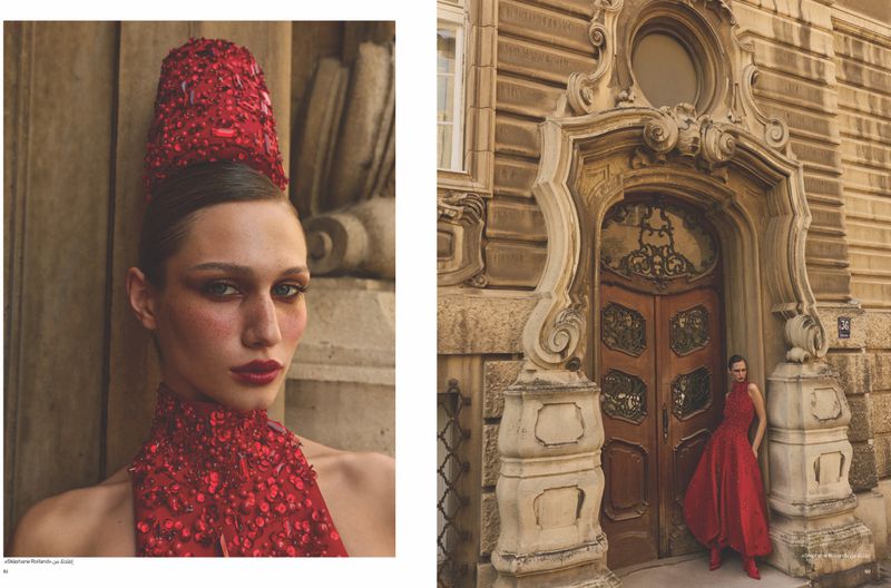 Close-up portrait of a woman wearing an elaborate red sequined dress and towering headpiece in front of carved classical stone architecture.