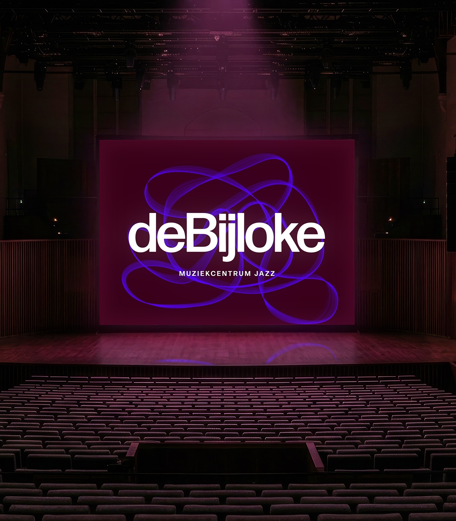 An illuminated concert hall featuring rows of empty seats facing a large stage screen displaying the de Bijloke brand identity in purple and white.