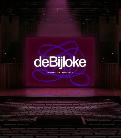 An illuminated concert hall featuring rows of empty seats facing a large stage screen displaying the de Bijloke brand identity in purple and white.