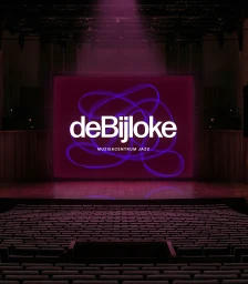 An illuminated concert hall featuring rows of empty seats facing a large stage screen displaying the de Bijloke brand identity in purple and white.